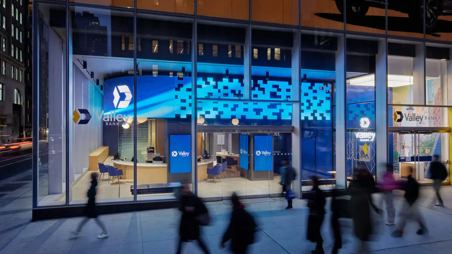 Modern Valley Bank storefront with large glass windows, illuminated blue digital signage, and people walking by on the sidewalk.