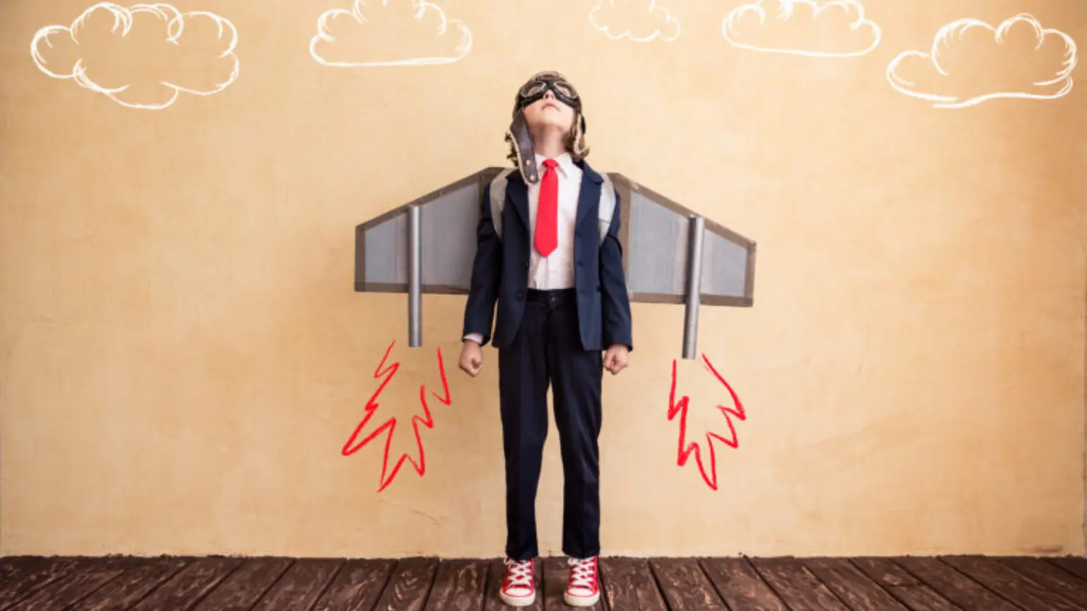 A child dressed in a business suit with a cardboard jetpack and drawn flames, looking upward.
