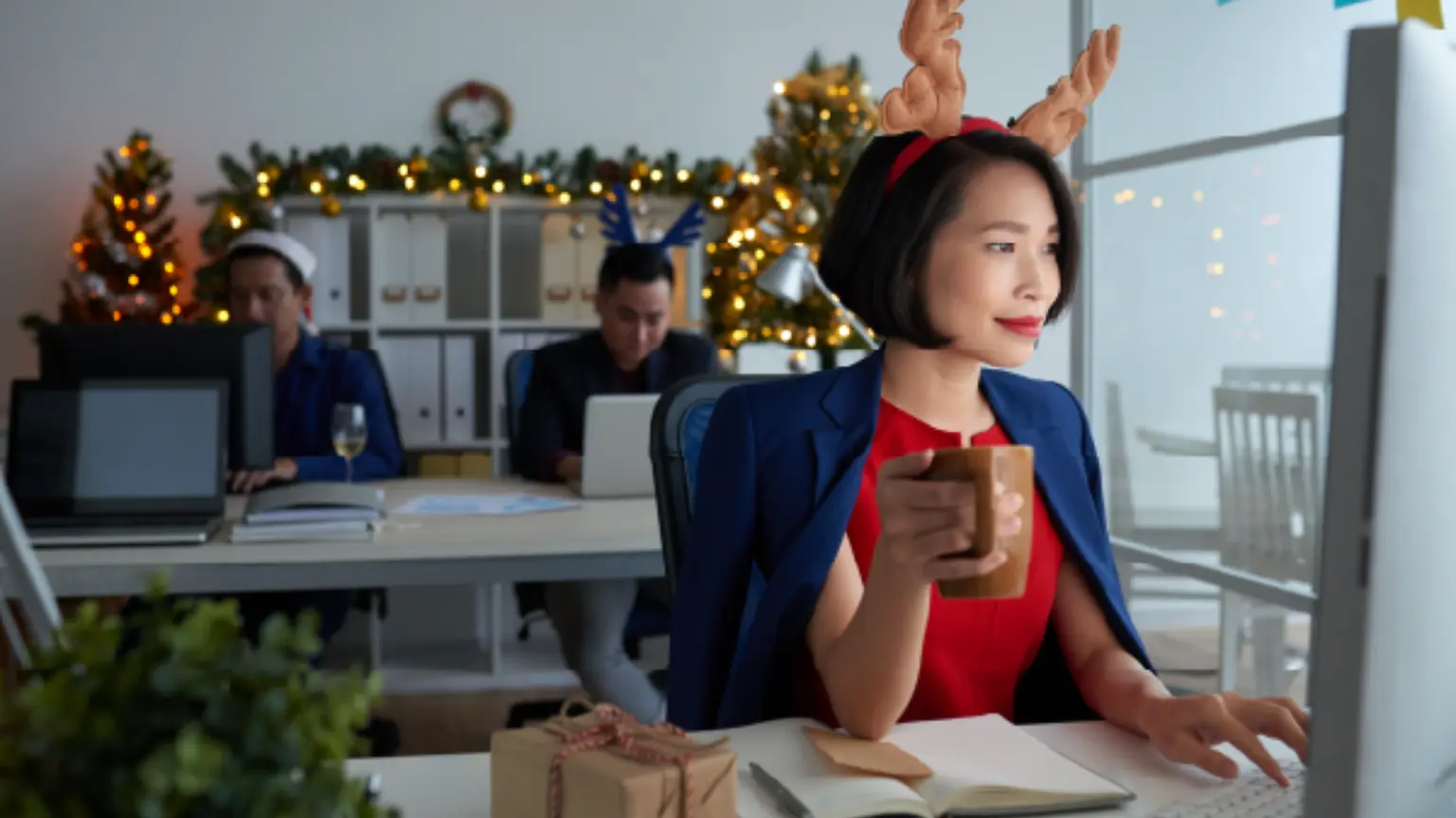 Woman in antler headband working at a desk with festive decorations and colleagues in background.