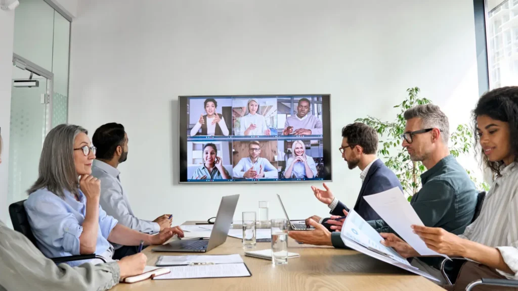 Office team participating in a video conference with colleagues shown on a large screen.
