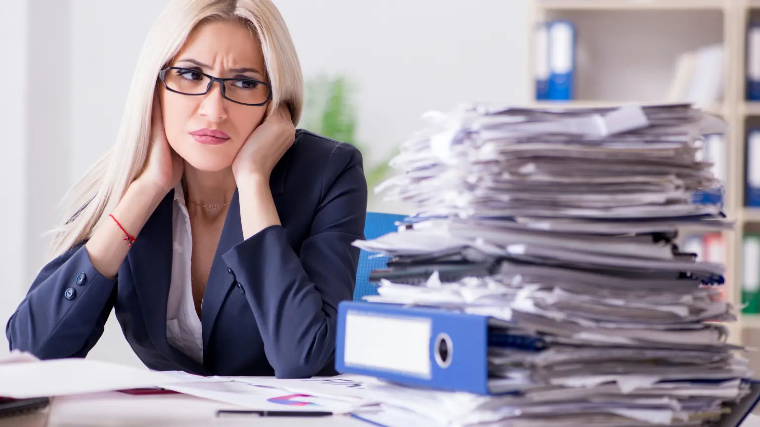 A frustrated businesswoman sits beside a large, disorganized stack of paperwork.