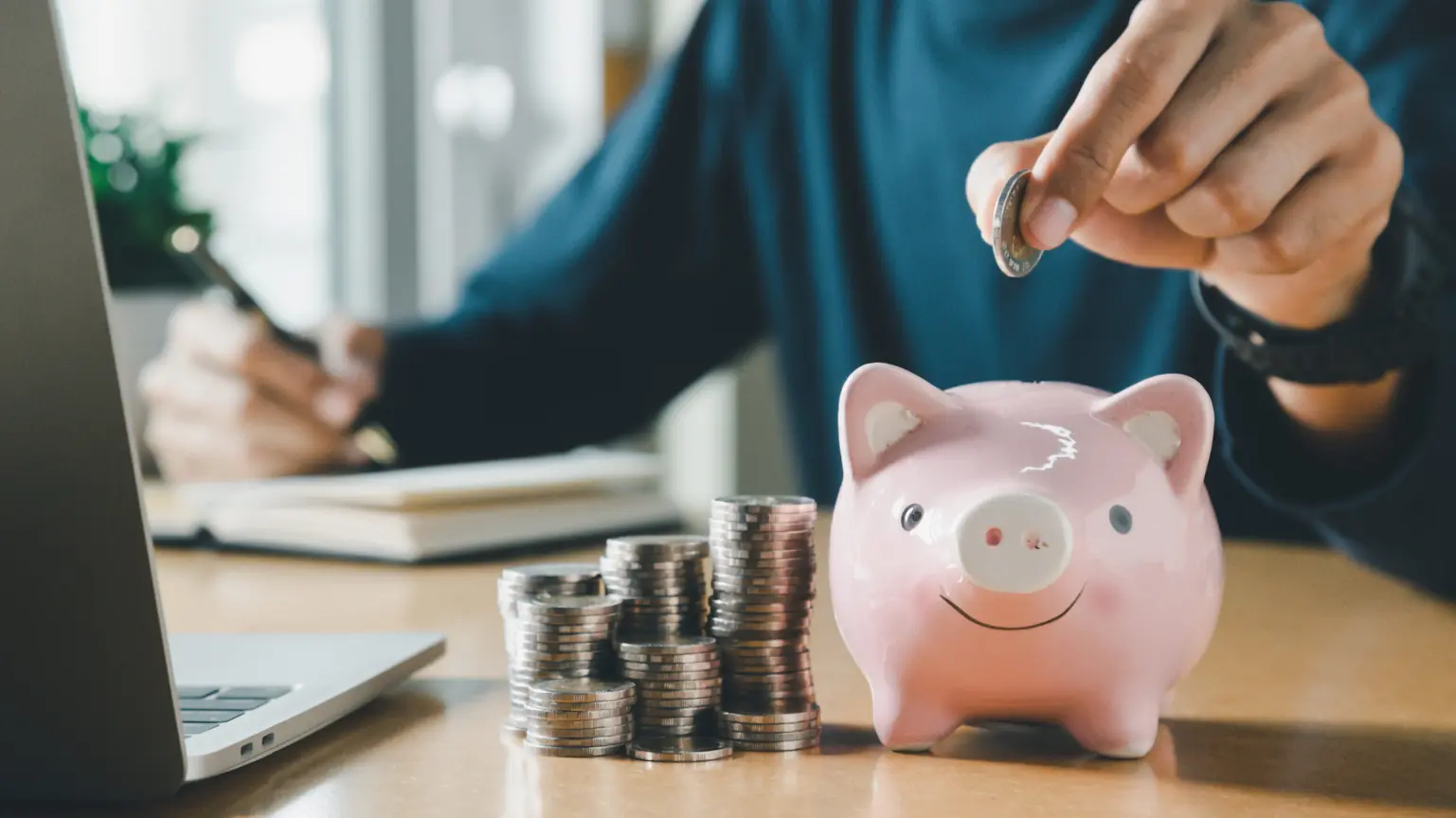 Person dropping a coin into a pink piggy bank next to stacks of coins on a desk.