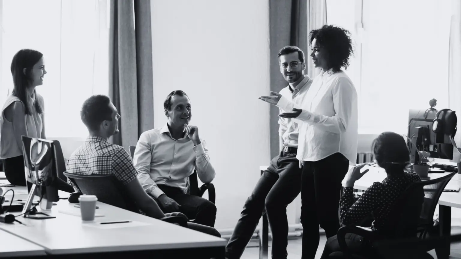 Black and white photo of a diverse group having a casual conversation in a modern office.