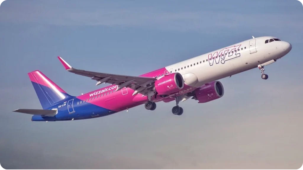 Wizz Air passenger airplane flying through a cloudy blue sky with its landing gear extended.