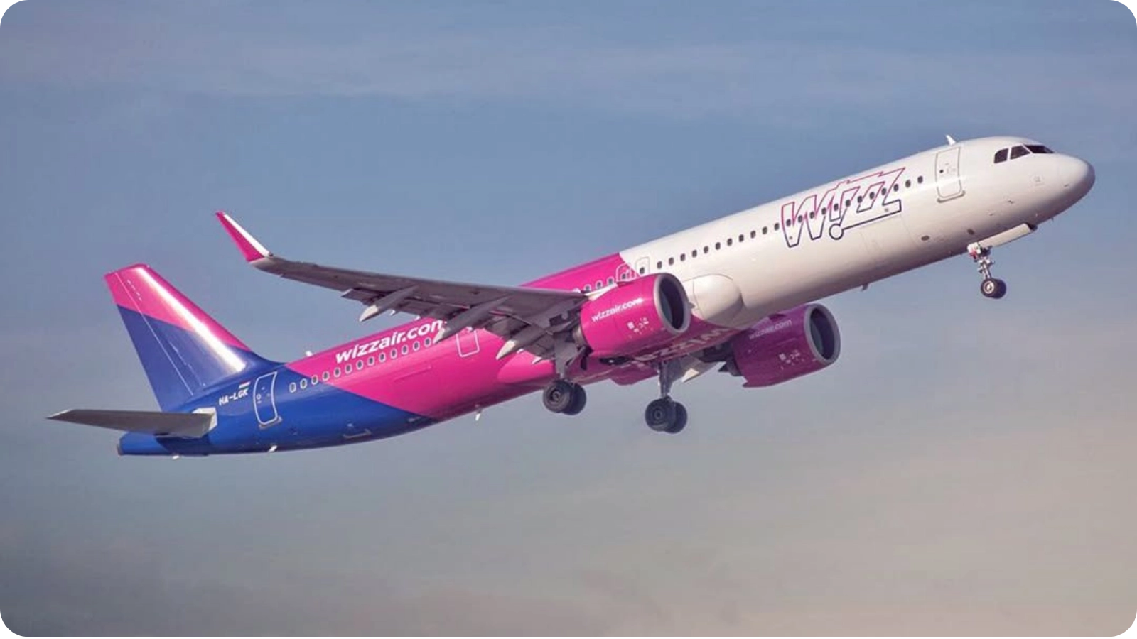Wizz Air passenger airplane flying through a cloudy blue sky with its landing gear extended.
